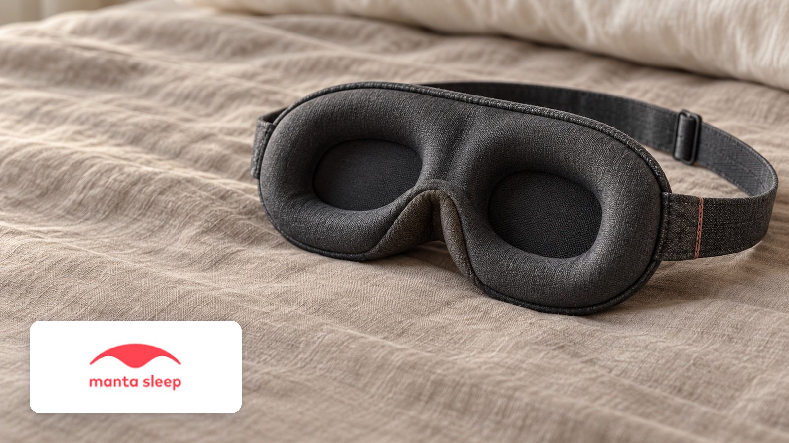 Manta Sleep logo with Charcoal contoured sleep mask with recessed eye cups and adjustable strap on linen bed